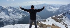 A man stands on a snowy mountain peak with arms raised, embracing the breathtaking view.